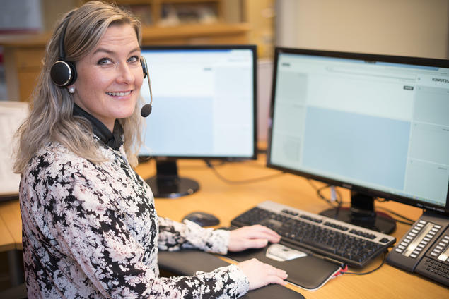 En kvinnelig resepsjonist med headset som smiler mot kameraet mens hun sitter ved en pult med to dataskjermer, tastatur og telefon. Foto.