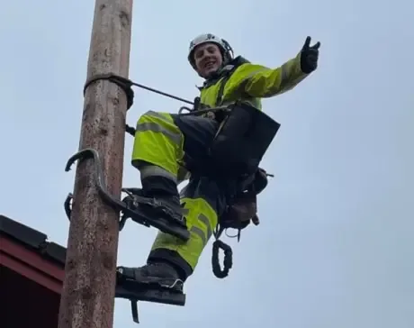 En lærling kledd i refleksjakke og hjelm klatrer i en stolpe ved hjelp av stigetrinn, smiler og gir tommel opp.En lærling kledd i refleksjakke og hjelm klatrer i en stolpe ved hjelp av stigetrinn, smiler og gir tommel opp.